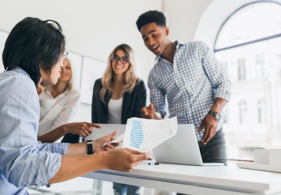 Asian manager in wristwatch sitting at his workplace and holding paper with graph. Indoor portrait of african programmer in checkered shirt talking with other employees in office.