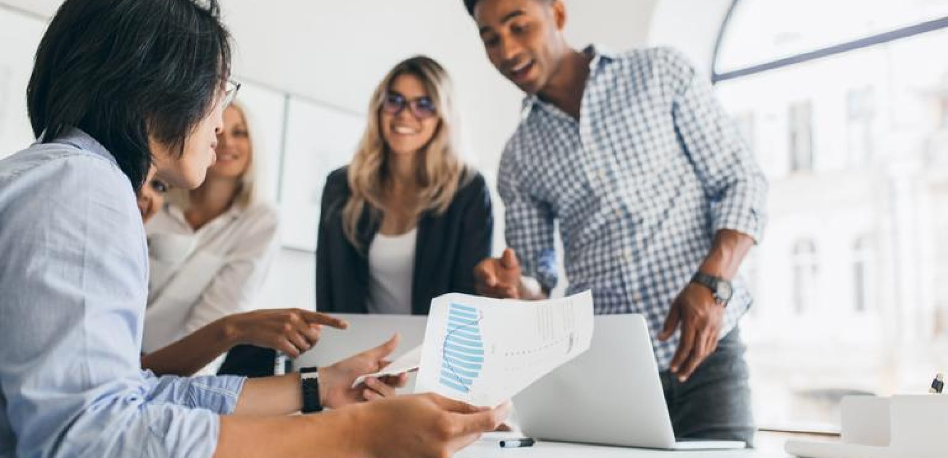 Asian manager in wristwatch sitting at his workplace and holding paper with graph. Indoor portrait of african programmer in checkered shirt talking with other employees in office.